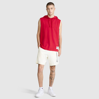 A young man stands against a plain light background, sporting a red Champion Jersey Sleeveless Hoodie with the C Logo and white shorts featuring the Champion text. He completes the look with white sneakers and displays tattoos on his left leg and right forearm.