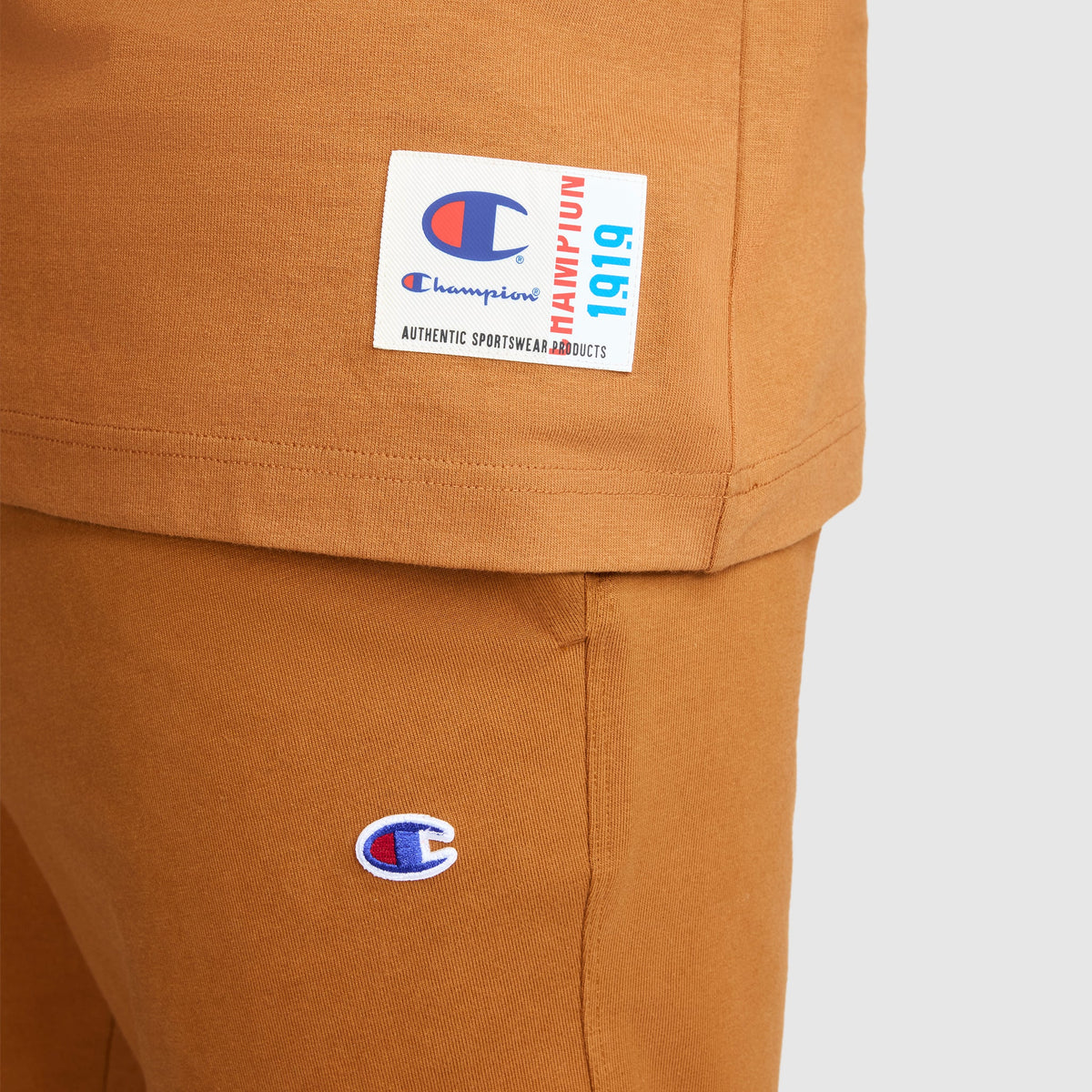 Close-up of a person wearing a matching brown Champion Jersey Sleeveless Hoodie with the C Logo, made from soft ring-spun cotton. The hoodie features the iconic Champion logo and a white label with blue and red text.