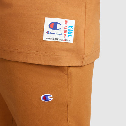 Close-up of a person wearing a matching brown Champion Jersey Sleeveless Hoodie with the C Logo, made from soft ring-spun cotton. The hoodie features the iconic Champion logo and a white label with blue and red text.