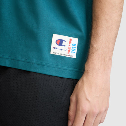 A close-up of a person’s right hand resting by their side, wearing the Champion Short Sleeve Boxy T-Shirt with an embroidered script logo—crafted from soft ring-spun cotton—paired with black mesh shorts and featuring a visible logo tag.