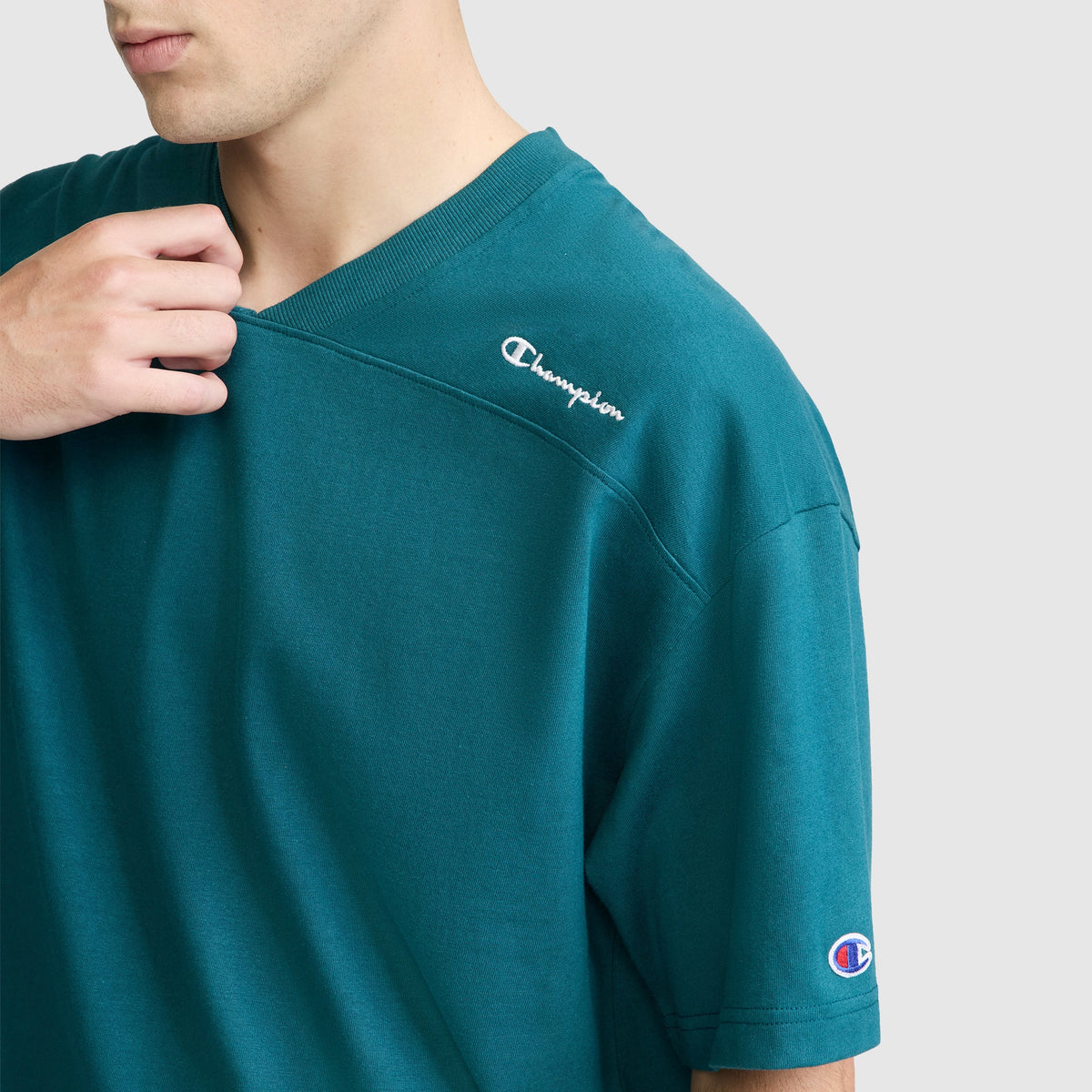 A person wearing a teal Champion Short Sleeve Boxy T-Shirt with an embroidered script logo gently holds the neckline. The Champion branding is visible on the chest and sleeve of this relaxed fit tee. The background is plain and light-colored.