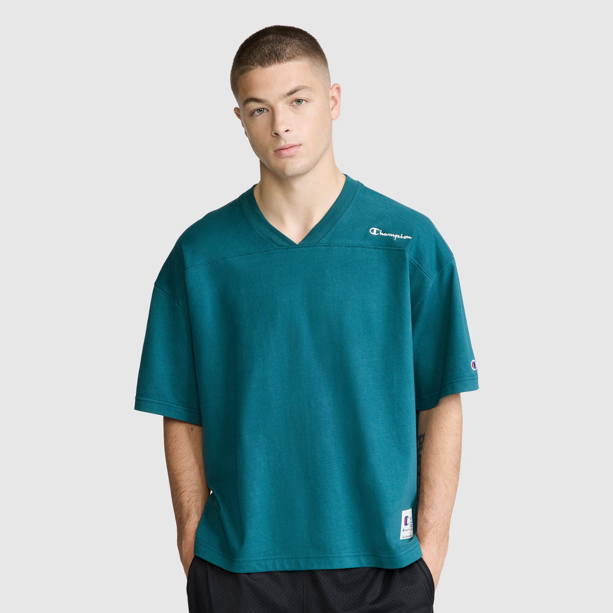 A person wearing a loose, teal Champion Short Sleeve Boxy T-Shirt with the embroidered script logo, made from ring-spun cotton, and black shorts stands against a plain light background, looking straight ahead with hands in pockets.