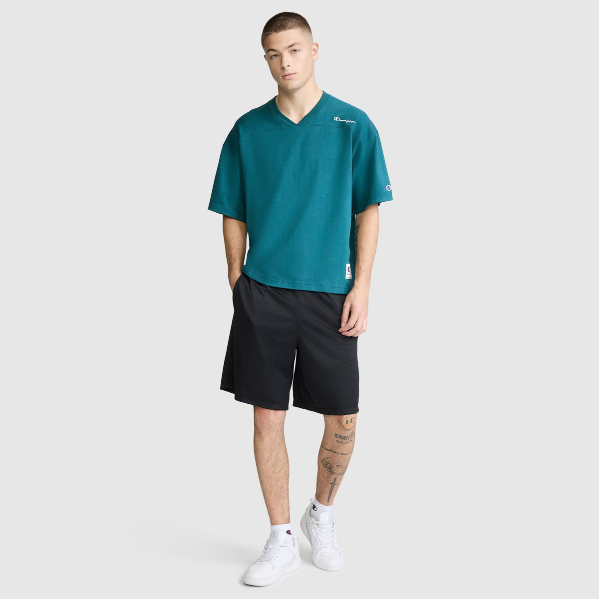 A young man is wearing the Champion Short Sleeve Boxy T-Shirt with an embroidered script logo, featuring a relaxed fit in teal ring-spun cotton. He pairs it with black shorts and white sneakers, displaying tattoos on his left leg as he poses with his left hand in his pocket against a plain light background.