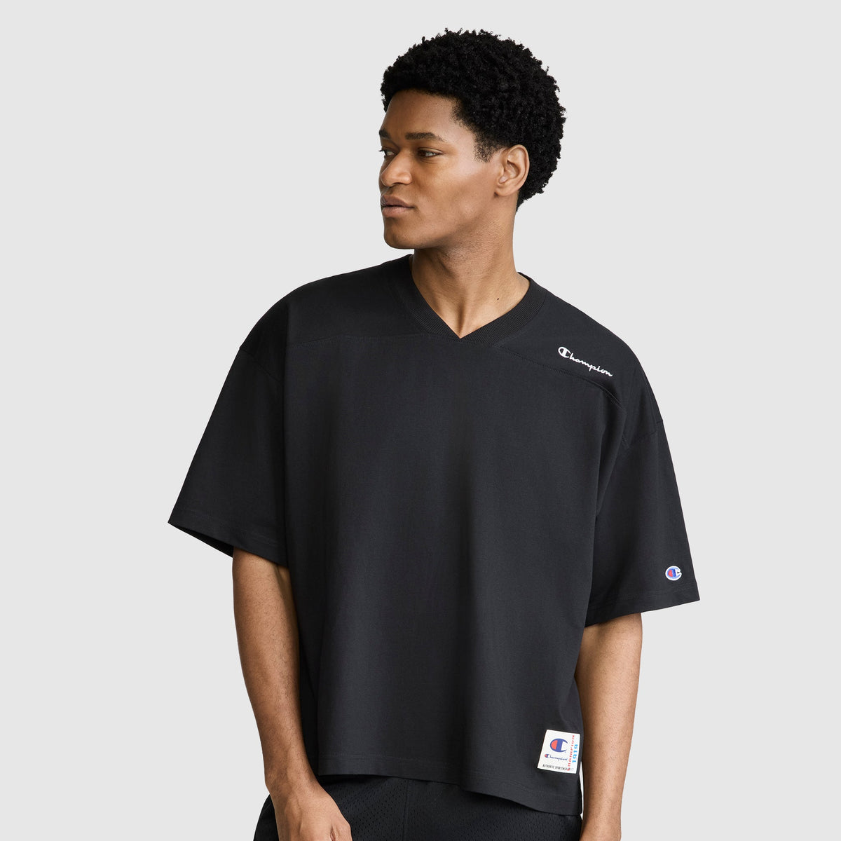 A man wearing a relaxed fit Champion Short Sleeve Boxy T-Shirt with an embroidered script logo—a loose black V-neck tee with short sleeves—stands against a light gray background, looking to his left with a neutral expression.