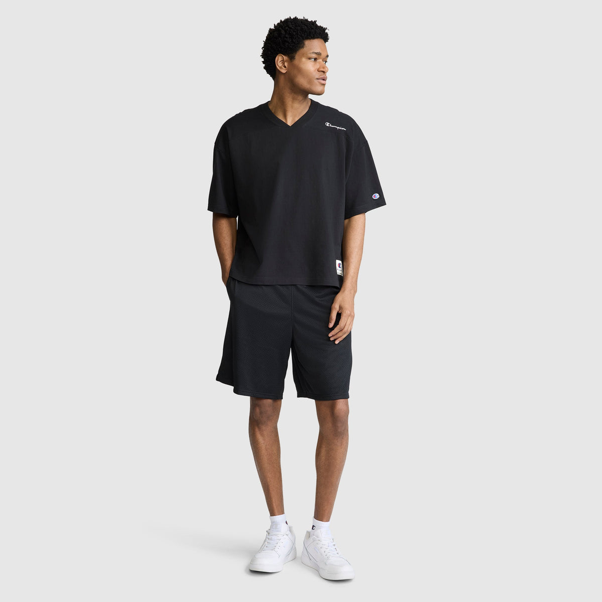 A man stands against a plain light gray background, wearing the Champion Short Sleeve Boxy T-Shirt with an embroidered script logo. The relaxed, loose black V-neck shirt is paired with black shorts and white sneakers. With his hands in his pockets and looking to his left, he exudes effortless heritage style.
