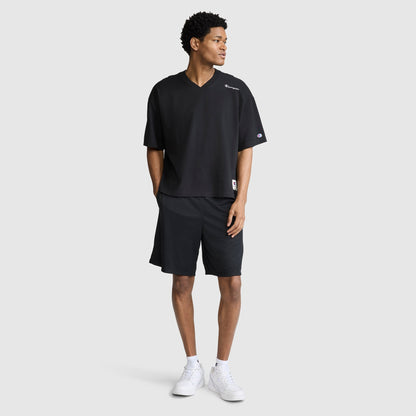 A man stands against a plain light gray background, wearing the Champion Short Sleeve Boxy T-Shirt with an embroidered script logo. The relaxed, loose black V-neck shirt is paired with black shorts and white sneakers. With his hands in his pockets and looking to his left, he exudes effortless heritage style.