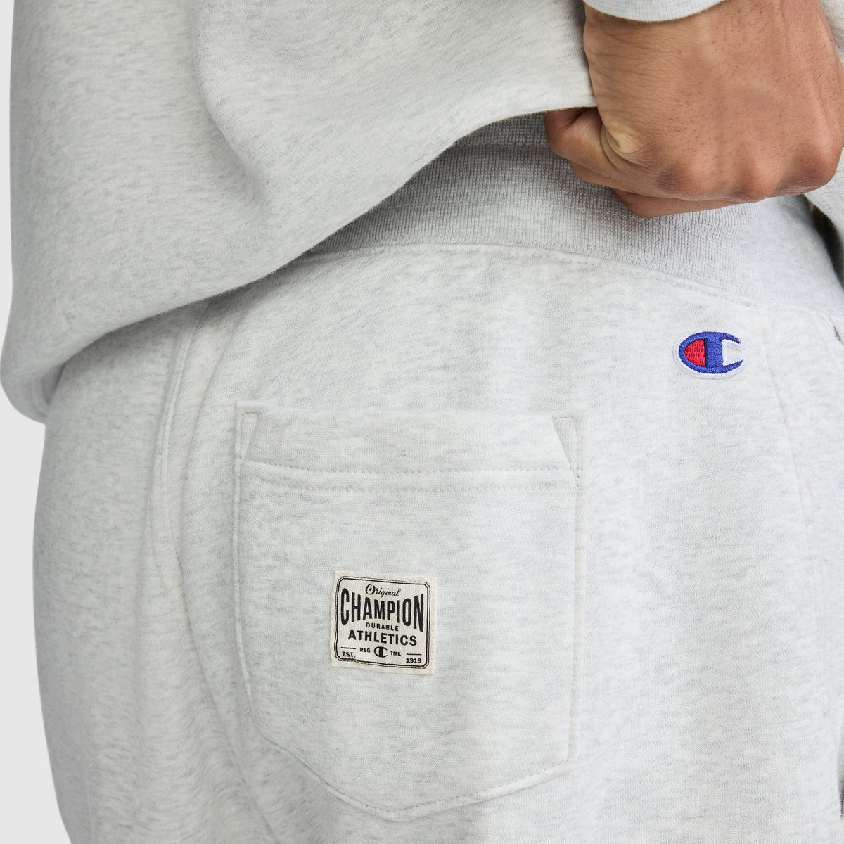 A close-up showcases a person wearing light gray Champion All in a Day’s Work Sweatpants. The photo highlights the back pocket with its Champion Athletics label and embroidered Champion logo, as a hand rests near the waistband—perfect fleece sweatpants for an easygoing, workwear-inspired look.