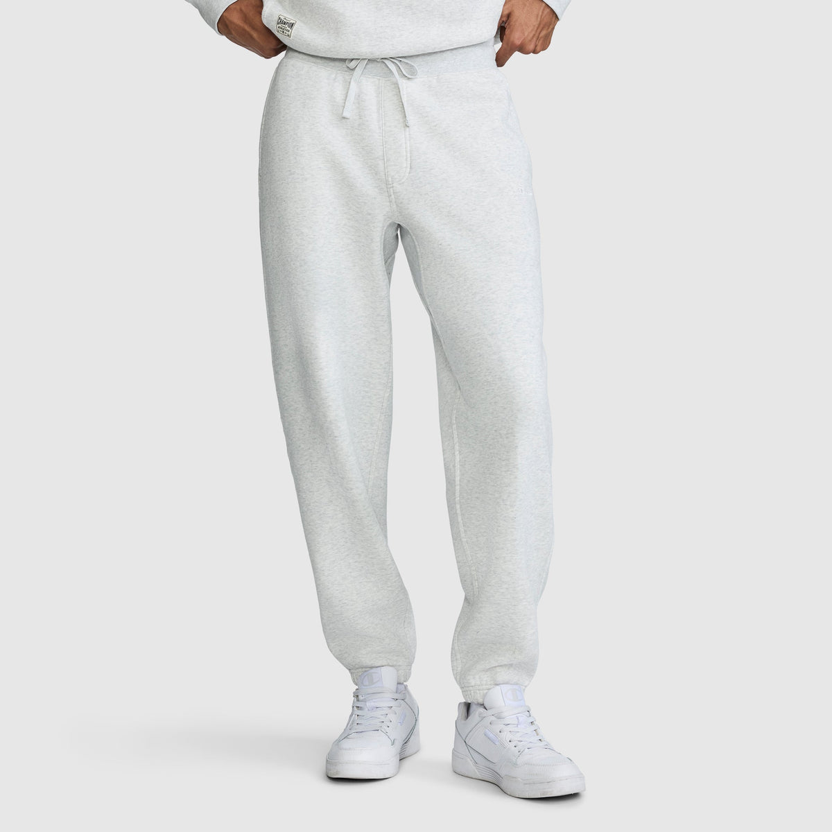 A person wearing Champion All in a Day's Work Sweatpants in light gray with a drawstring, paired with white sneakers. The individual stands with hands at their waist, and the upper body is partially visible, showing a matching sweatshirt. The background is plain white.