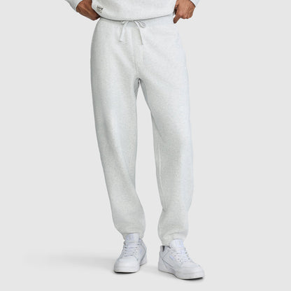 A person wearing Champion All in a Day's Work Sweatpants in light gray with a drawstring, paired with white sneakers. The individual stands with hands at their waist, and the upper body is partially visible, showing a matching sweatshirt. The background is plain white.