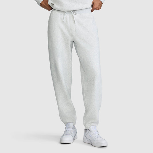 A person wearing Champion All in a Day's Work Sweatpants in light gray with a drawstring, paired with white sneakers. The individual stands with hands at their waist, and the upper body is partially visible, showing a matching sweatshirt. The background is plain white.