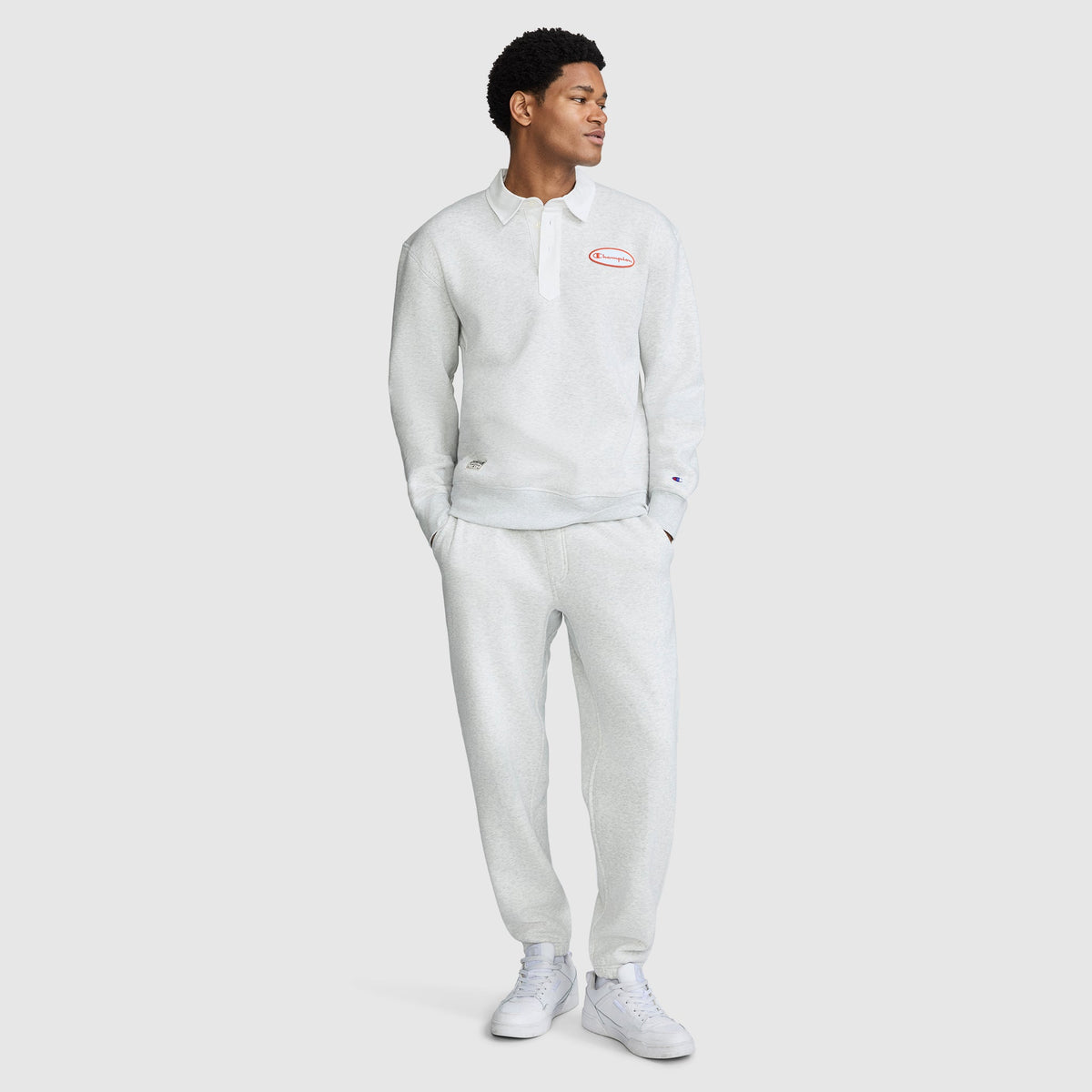 A person stands wearing a light gray long-sleeve collared shirt and the Champion All in a Day's Work Sweatpants, paired with white sneakers. The shirt features a small red and white patch on the chest.