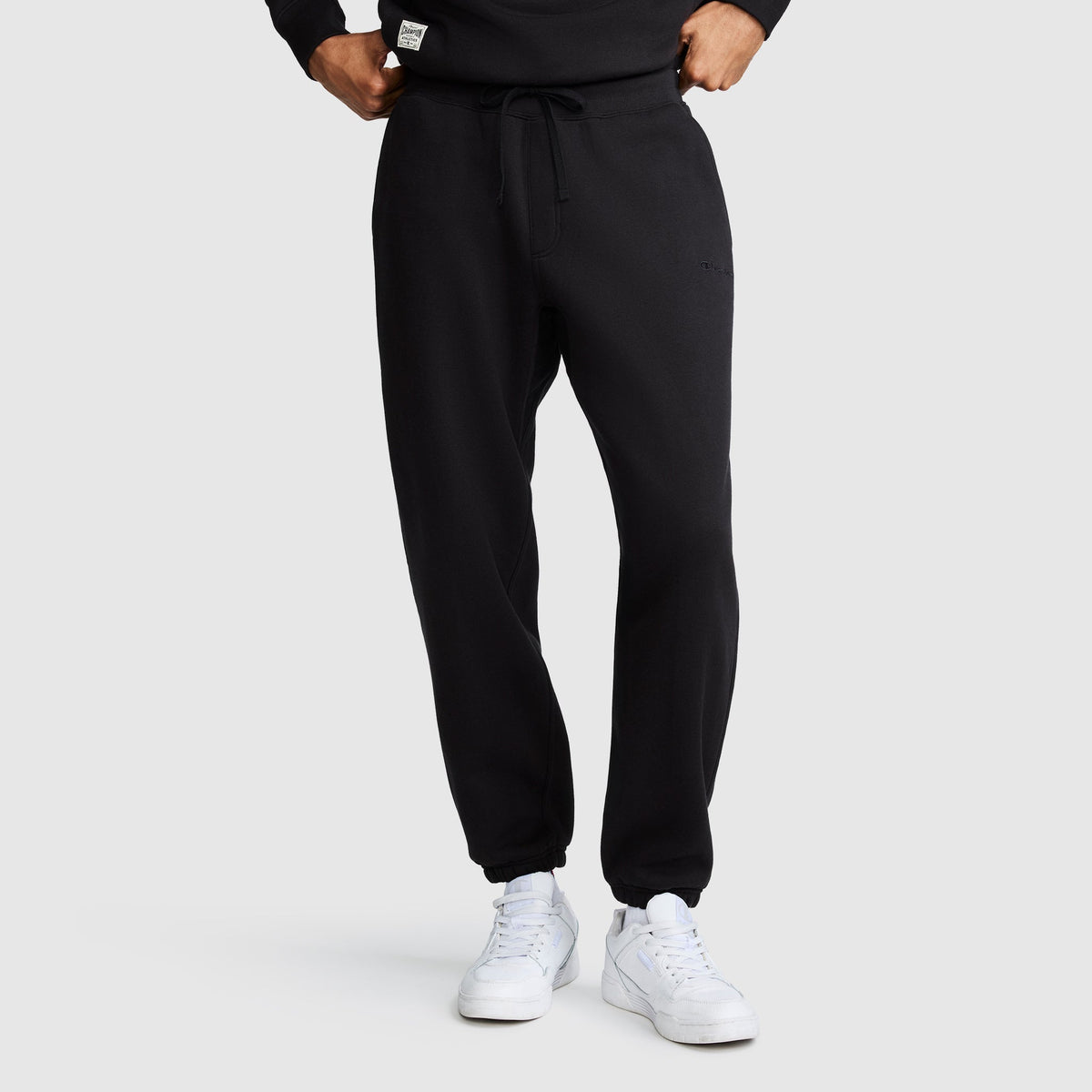 A person is shown from the waist down, standing against a plain light gray background, wearing Champion’s All in a Day's Work Sweatpants in black and white sneakers. Their hands rest casually in or near the pockets of the fleece pants.