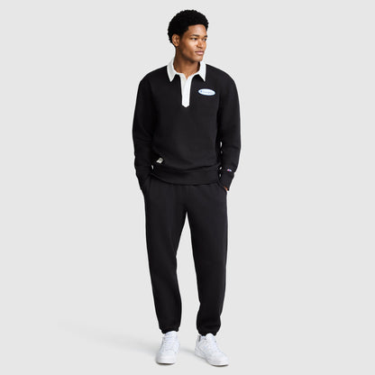 A man stands facing slightly left, wearing a black collared sweatshirt with a white patch, All in a Day's Work Sweatpants by Champion in black, and white sneakers, against a plain light gray background.