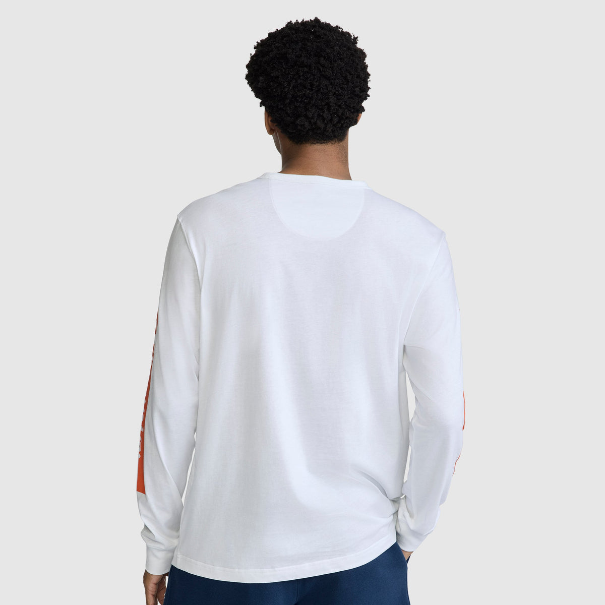 A person with short curly hair is wearing the Champion Medallion Long Sleeve Graphic T-Shirt and dark pants, standing with their back to the camera against a plain light gray background.