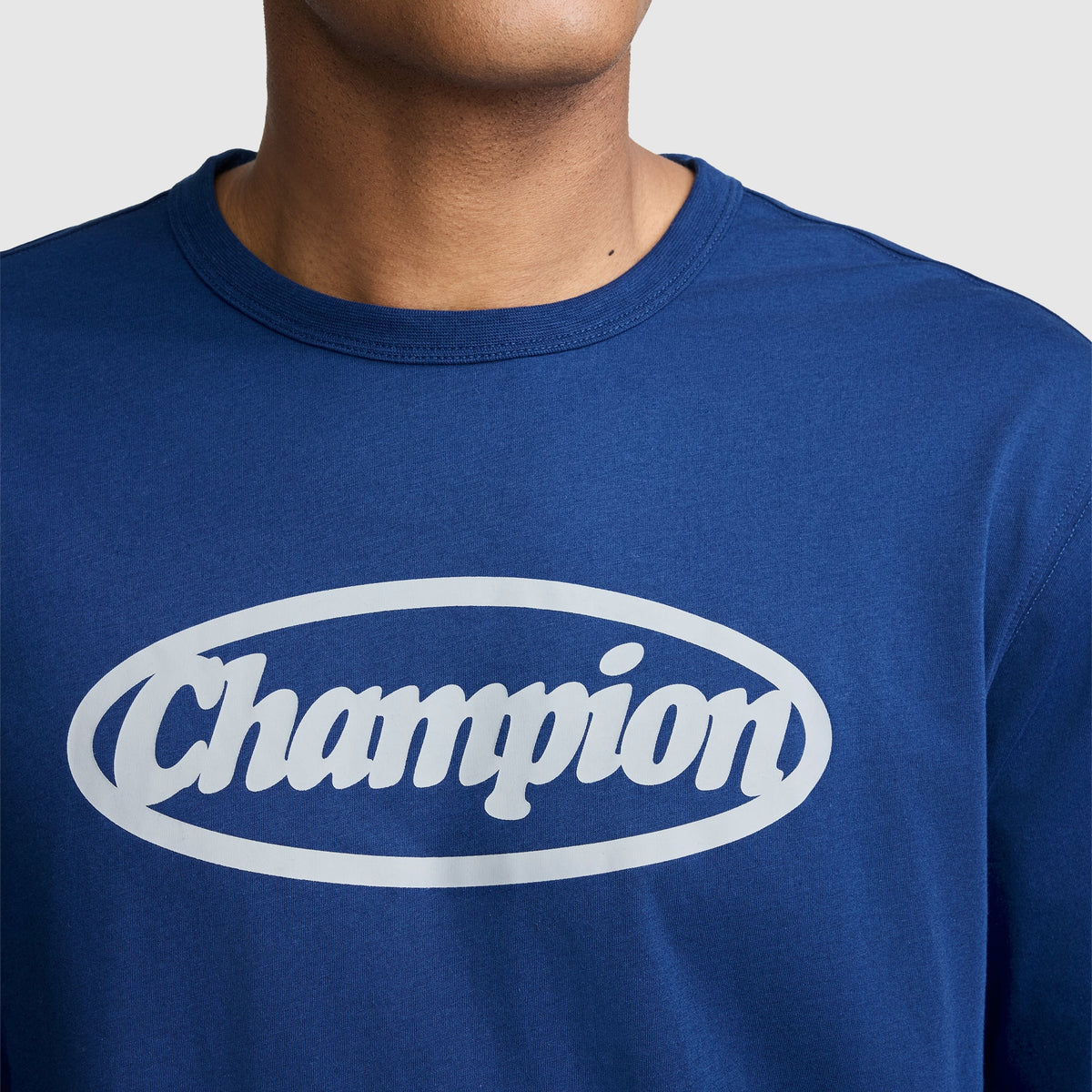 A person wearing a blue Champion Medallion Long Sleeve Graphic T-Shirt, featuring the word “Champion” in white inside an oval on the chest. Only the lower half of their face and neck are visible, subtly referencing moto sport heritage style.