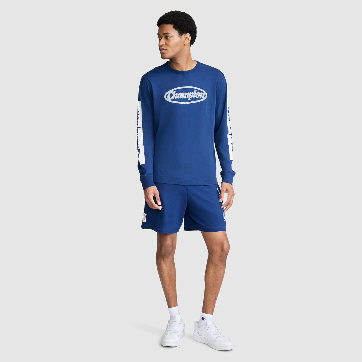 A man wearing a blue Champion Medallion Long Sleeve Graphic T-Shirt and matching shorts stands facing slightly to the right, channeling motorsport heritage style. He has short hair, is wearing white sneakers, and stands against a plain light gray background.