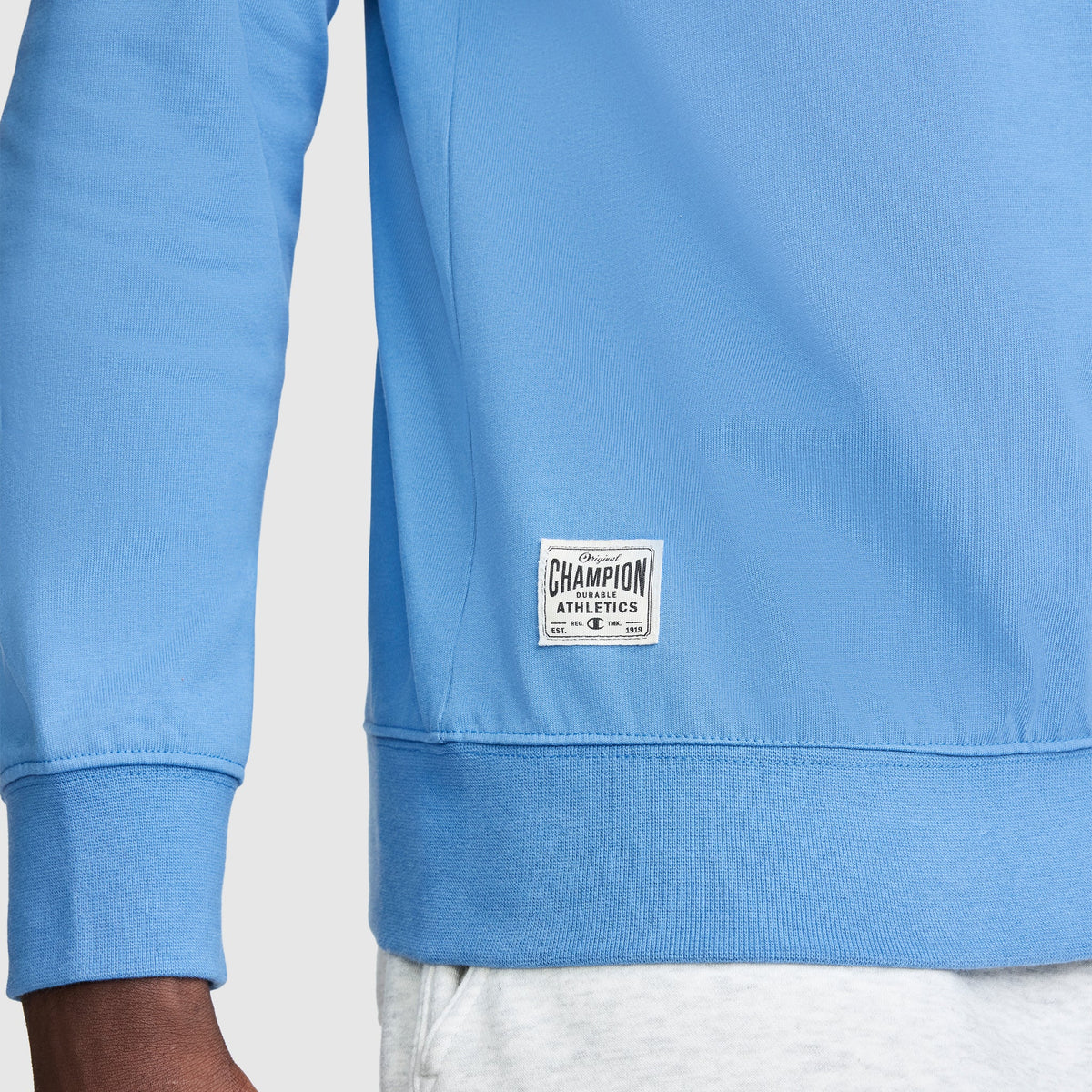 A person wearing a light blue Champion Striped Rugby Shirt in an athletic style and white pants. Only the lower part of the long sleeve shirt, left arm, and part of the hand are visible.