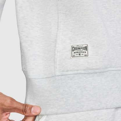 A close-up of a person wearing the Champion Fleece Rugby in light grey, featuring a small logo patch near the hem. The person is holding the hem of the Fleece Rugby with their left hand.
