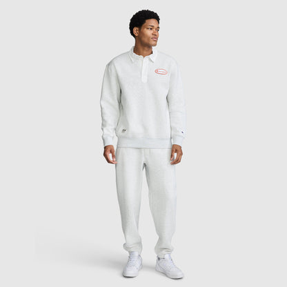 A person stands wearing a light gray Champion Fleece Rugby pullover and matching sweatpants with white sneakers. The Champion Fleece Rugby features a collar, buttons, and a red Champion logo on the chest. The background is plain and light.