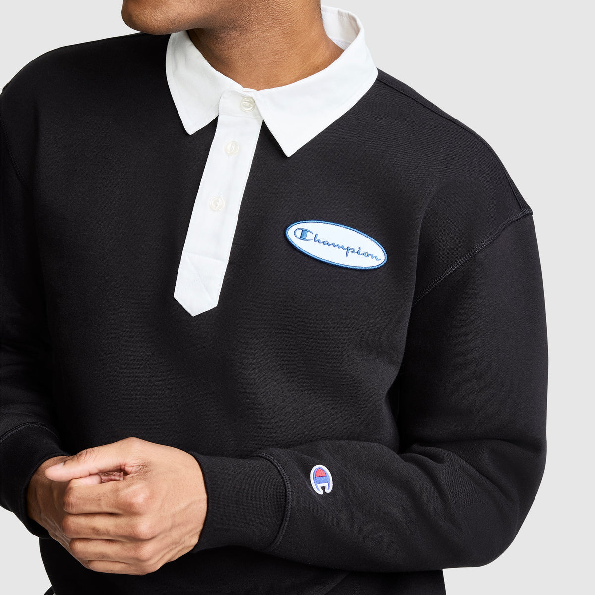 A person wearing the Champion Fleece Rugby, featuring a black design with a white collar, a Champion logo patch on the chest, and a small Champion logo on the sleeve, stands against a plain background.