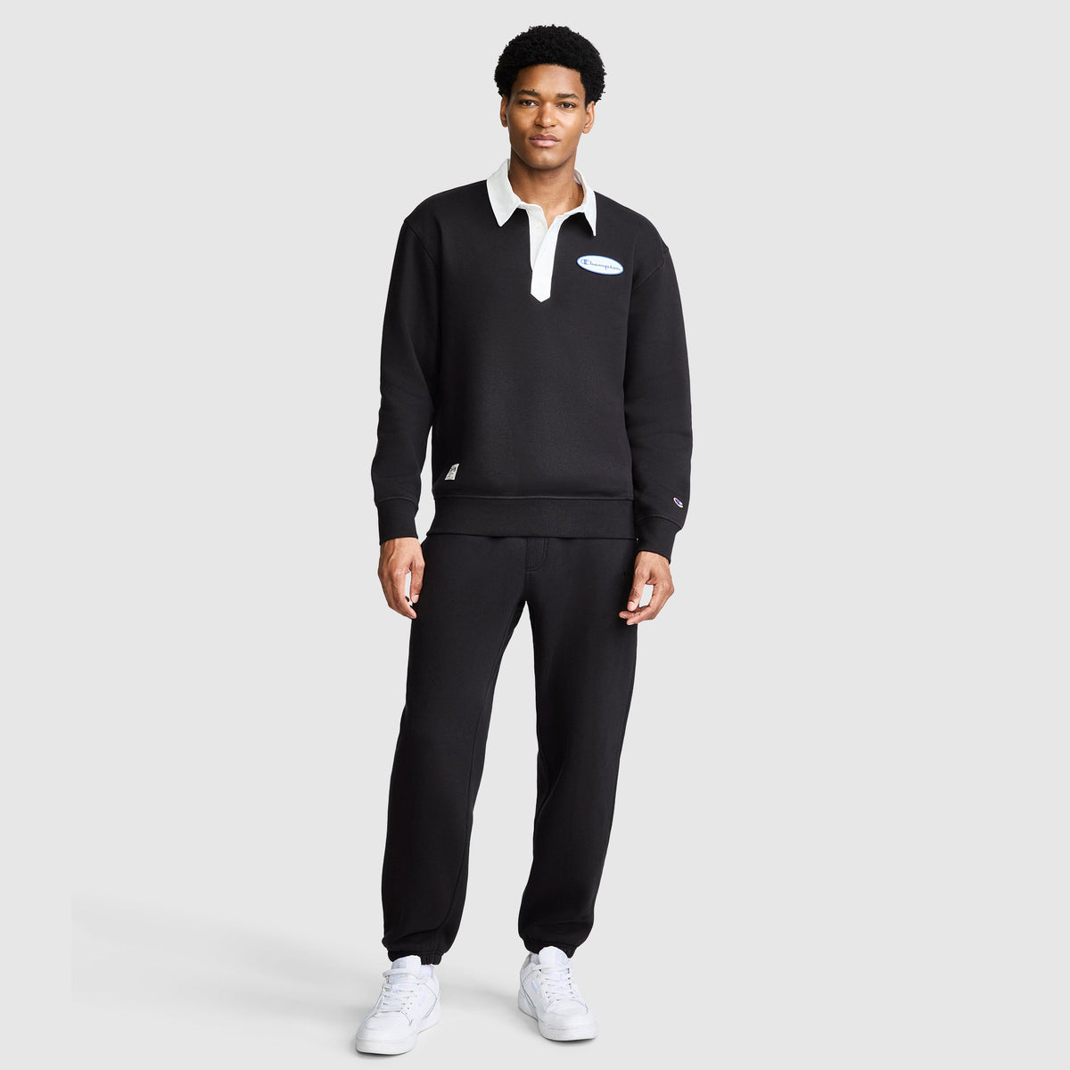 A man stands against a plain background wearing the Champion Fleece Rugby shirt with a white collar, paired with matching black pants and white sneakers. He faces forward with a neutral expression.