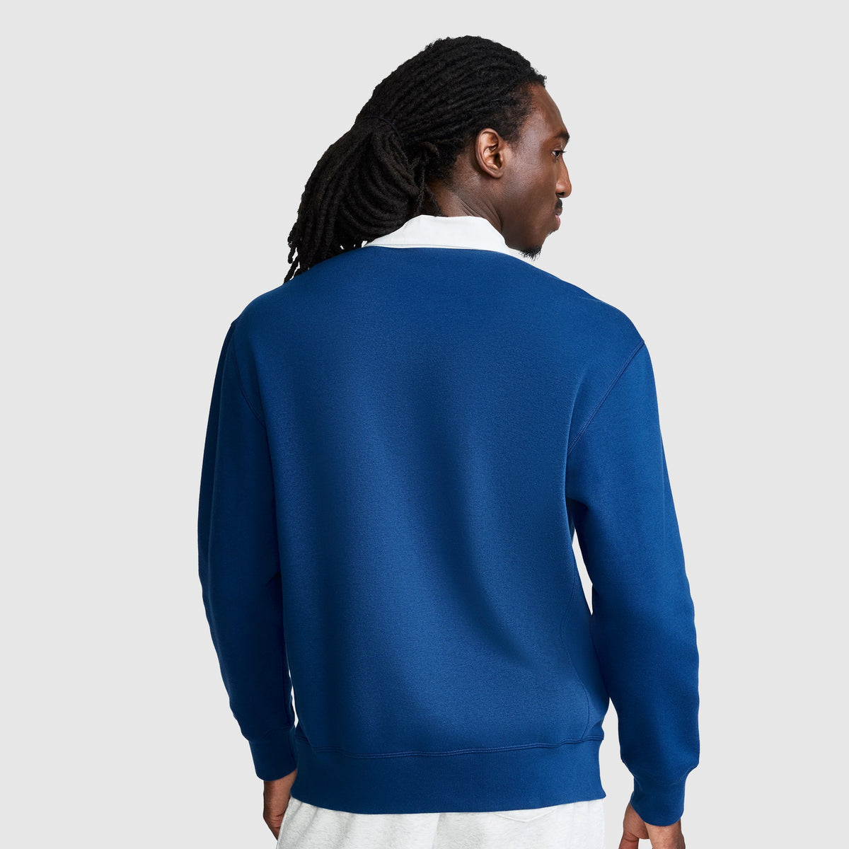 A person with long, dark hair in dreadlocks is shown from the back, wearing the Champion Fleece Rugby shirt in blue layered over a white shirt against a light gray background.