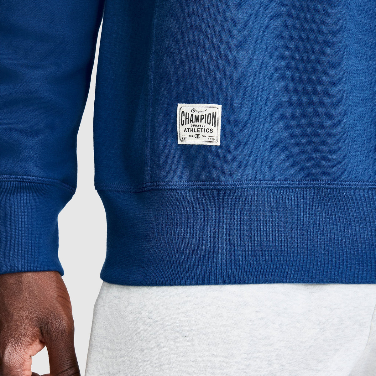 A close-up of a person wearing a blue Champion Fleece Rugby sweatshirt and light gray pants. Only part of the torso and the left hand are visible. The Champion label, sewn near the hem, highlights this classic Fleece Rugby style.