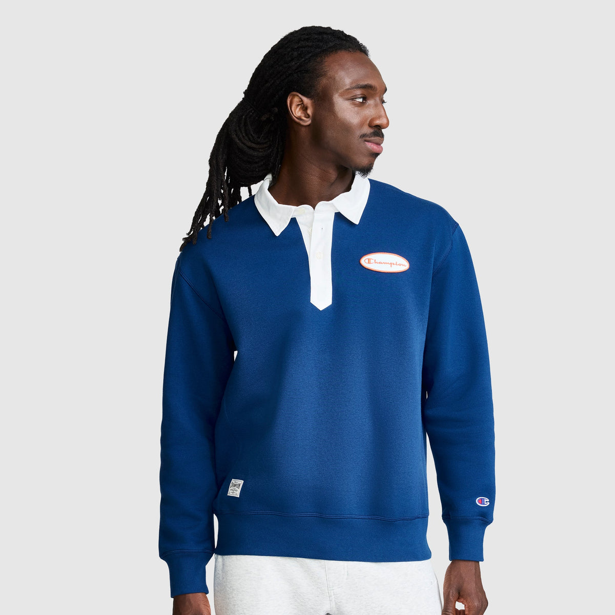 A person wearing the Champion Fleece Rugby shirt in blue, featuring a white collar, along with gray pants, stands against a plain light background and looks to the side.