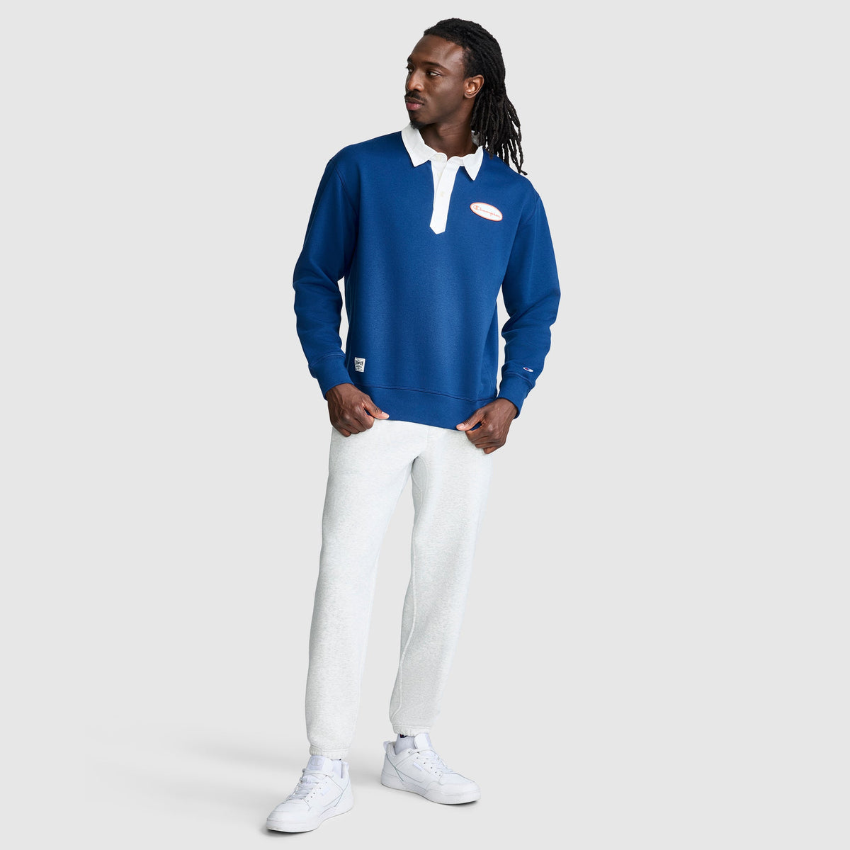 A man stands against a plain background, wearing the Champion Fleece Rugby shirt with a white collar and logo patch, paired with white pants and white sneakers. He looks to his left with one hand in his pocket.