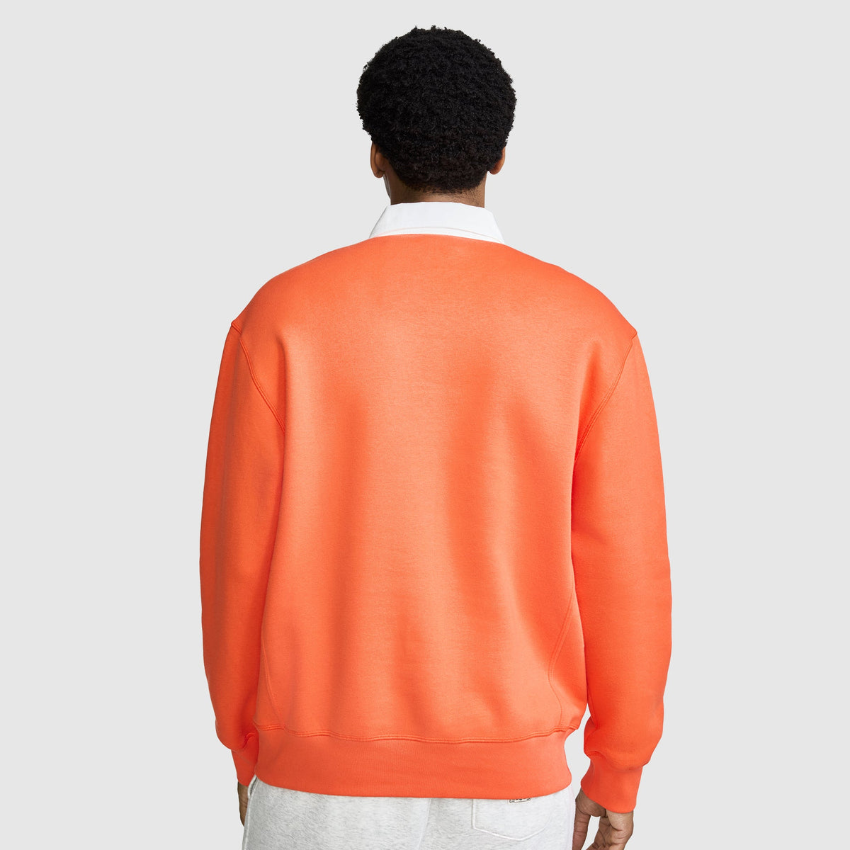 A person with short curly hair stands facing away against a plain light gray background, wearing the Champion Fleece Rugby in orange layered over a white collared shirt and paired with light gray pants.