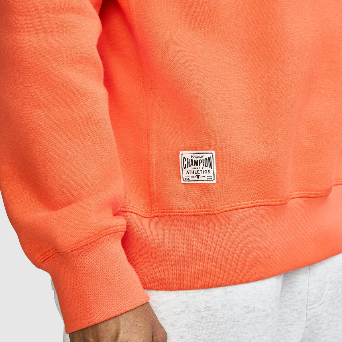 A person wears a bright orange Champion Fleece Rugby shirt and light gray pants. Only the torso and arm are visible, with a Champion logo label sewn near the hem of the shirt.