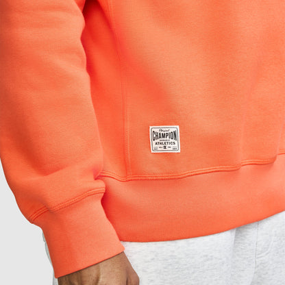 A person wears a bright orange Champion Fleece Rugby shirt and light gray pants. Only the torso and arm are visible, with a Champion logo label sewn near the hem of the shirt.