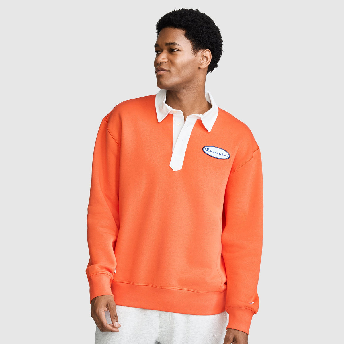 A man wearing the Champion Fleece Rugby, featuring an orange design with a white collar and a blue chest logo, stands against a light gray background, looking to the side.