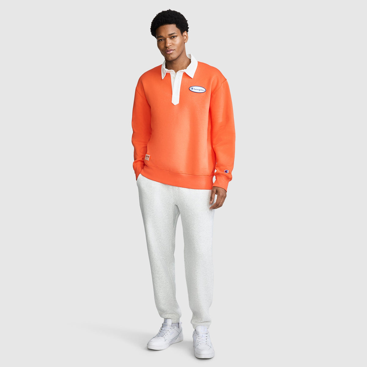 A man stands against a light background, wearing the Champion Fleece Rugby—a men's orange pullover with a white collar and patch—paired with light gray sweatpants and white sneakers. He faces forward with a relaxed expression.