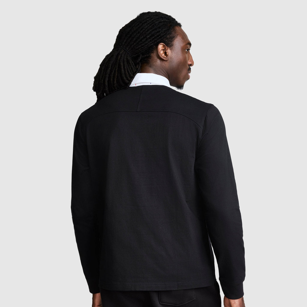A person with long, dark, braided hair facing away is wearing a black jacket over the Champion Long Sleeve Rugby Shirt with Twill Collar and Embroidered C Logo, featuring a white collar and long sleeves, set against a plain light gray background.