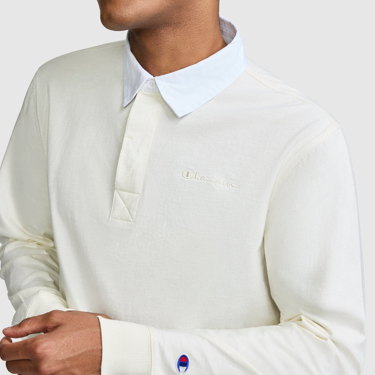 A person wearing the Champion Long Sleeve Rugby Shirt with Twill Collar and embroidered C logo, featuring cream-colored fabric and a classic white collar. The logos are visible on the chest and sleeve, while the person’s face is partially out of frame.