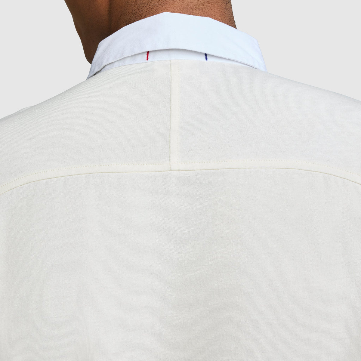 Close-up of the back of a person wearing a white Long Sleeve Rugby Shirt with Twill Collar from Champion, featuring a visible seam and subtle blue and red stitching near the collar, evoking classic rugby shirt details with an embroidered C logo.