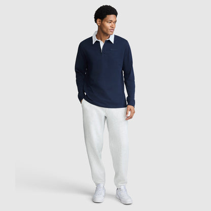 A man is wearing a navy Champion Long Sleeve Rugby Shirt with Twill Collar and Embroidered C Logo, paired with light gray sweatpants and white sneakers, standing and looking to the side against a plain light gray background.
