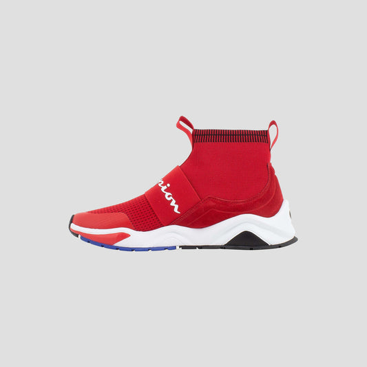 The Champion Kids' Rally Pro is a red high-top sneaker with a sock-like knit upper, white script on the side, and a chunky white/black sole. Pull tabs on the tongue and heel are shown, displayed on a light gray background.
