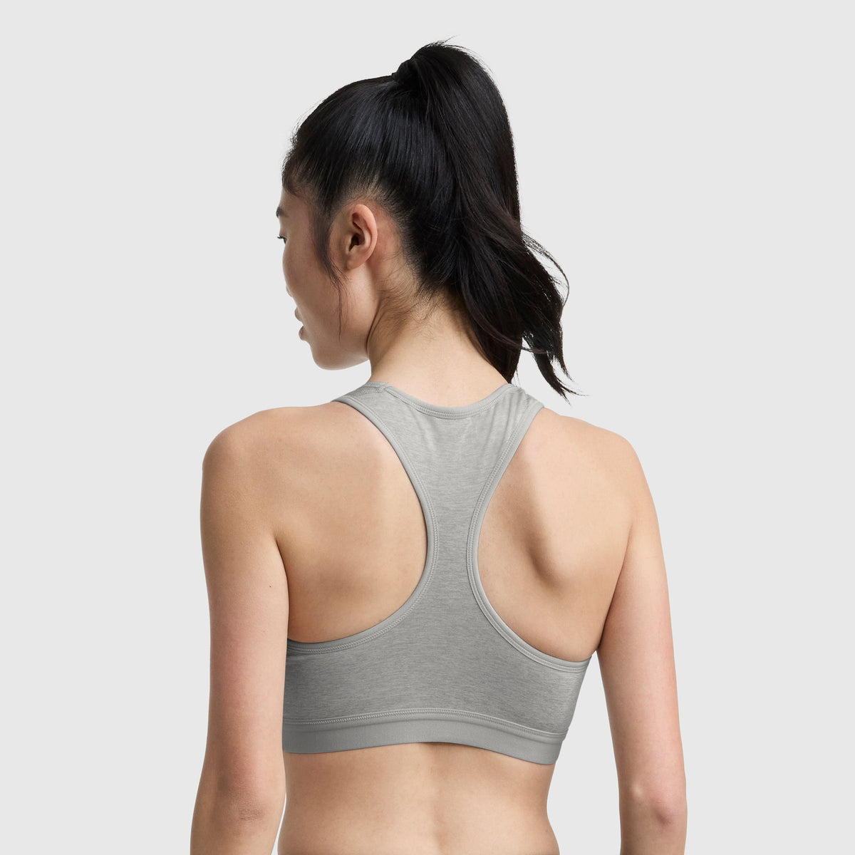 A woman with long dark hair in a ponytail is seen from behind wearing the Champion Compression Sports Bra, C Logo, in light gray against a plain light background.