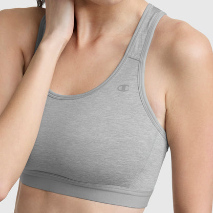 A woman is shown from shoulders to waist, wearing the Champion Compression Sports Bra with C Logo in light gray, offering moderate support. Her left hand touches her neck and the background is plain and light-colored.