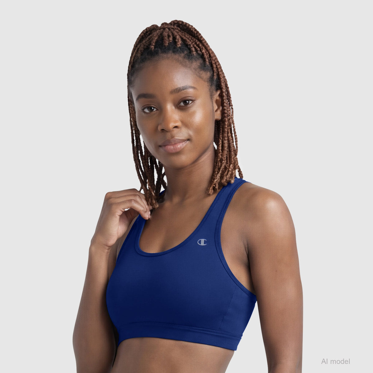 A woman with braided hair wears the Champion Compression Sports Bra, C Logo in blue, standing against a plain light background and looking at the camera with a slight smile.