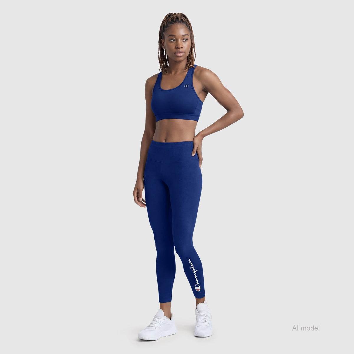 A woman models a blue Champion Compression Sports Bra, C Logo, and matching leggings with the Champion logo on the lower leg, paired with white sneakers against a plain gray background.