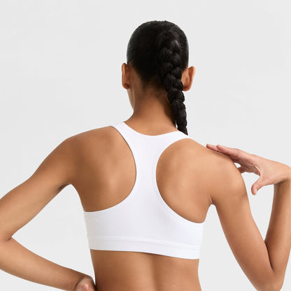 A woman with a long braided ponytail is seen from behind, raising her right arm near her shoulder while wearing the white Champion Compression Sports Bra, C Logo. The background is plain and light-colored.