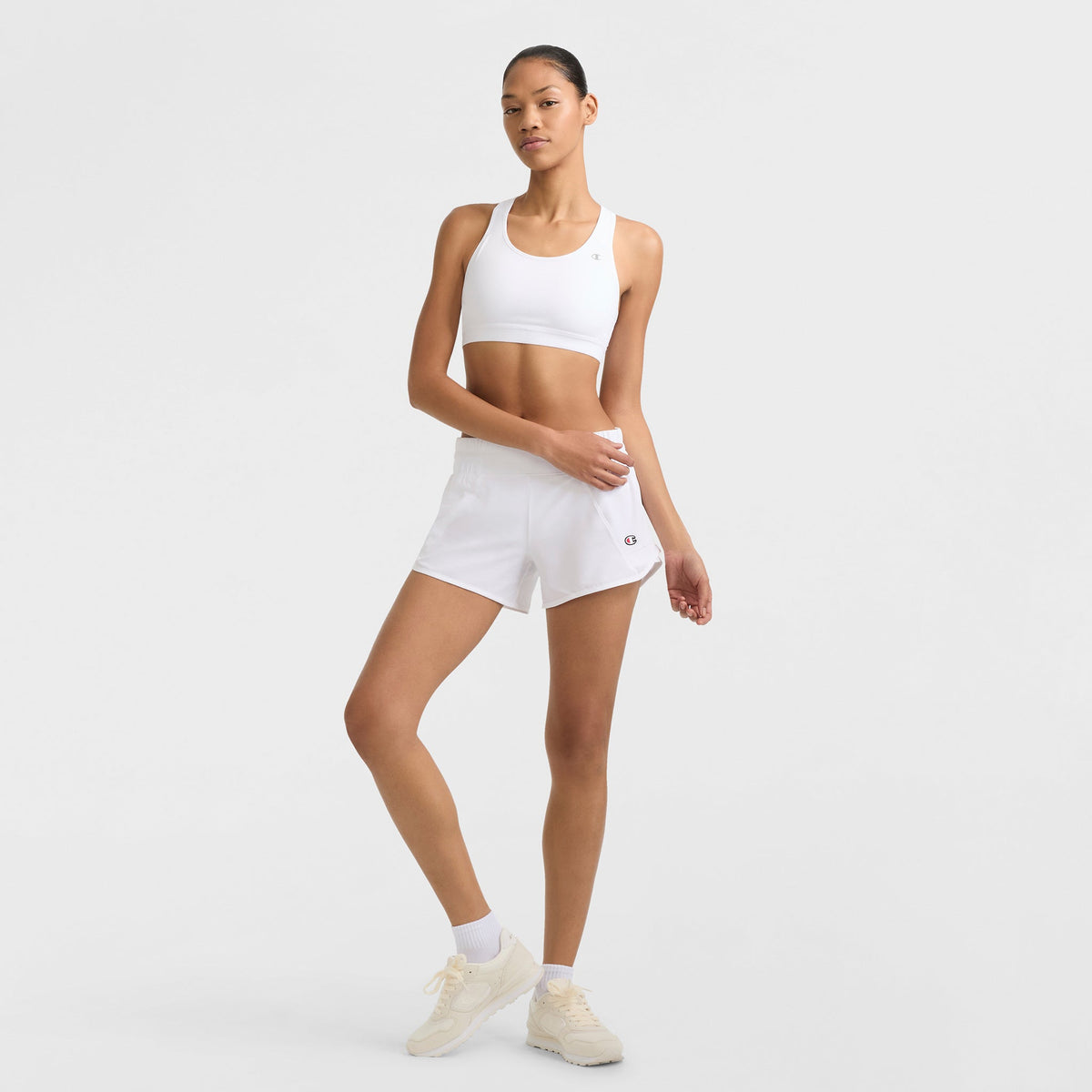 A woman poses against a plain background wearing a Champion Compression Sports Bra with C Logo, paired with white athletic shorts, white socks, and light sneakers. One hand rests on her waist while the other hangs by her side.