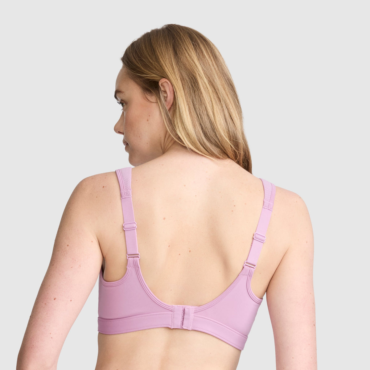 A woman with long blonde hair is shown from the back, wearing the Champion Spot Comfort High-Impact Sports Bra, C Logo in light purple with adjustable straps designed for maximum support against a plain light gray background.
