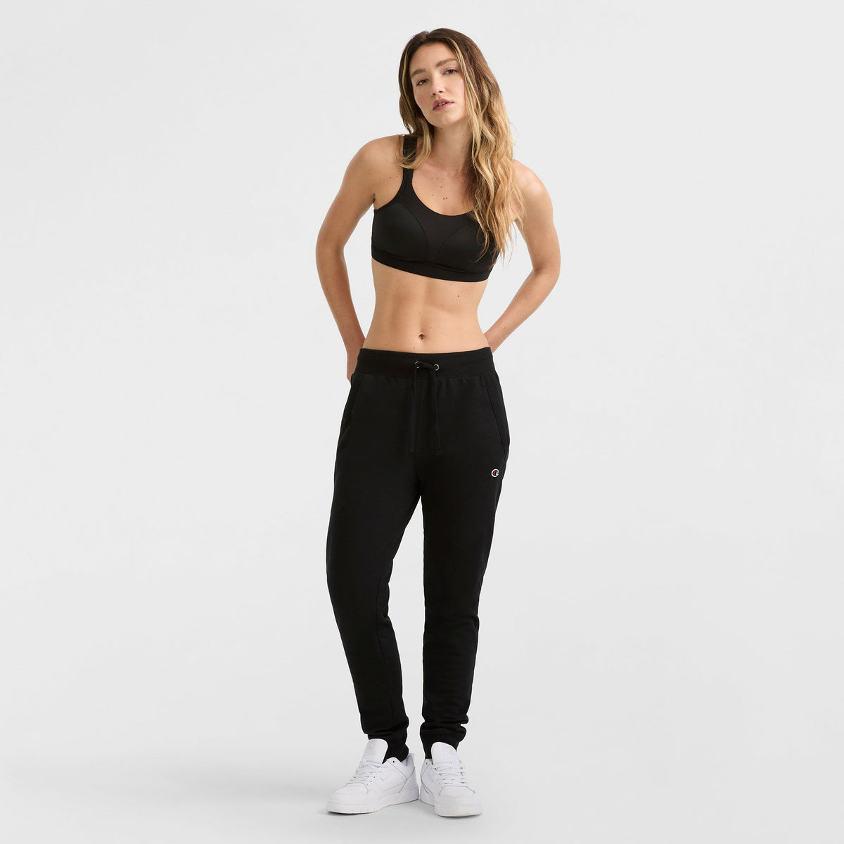 A woman with long, wavy hair stands against a white background, wearing the Champion Spot Comfort High-Impact Sports Bra with C Logo, paired with black jogger pants and white sneakers. Her hands are in her pockets.