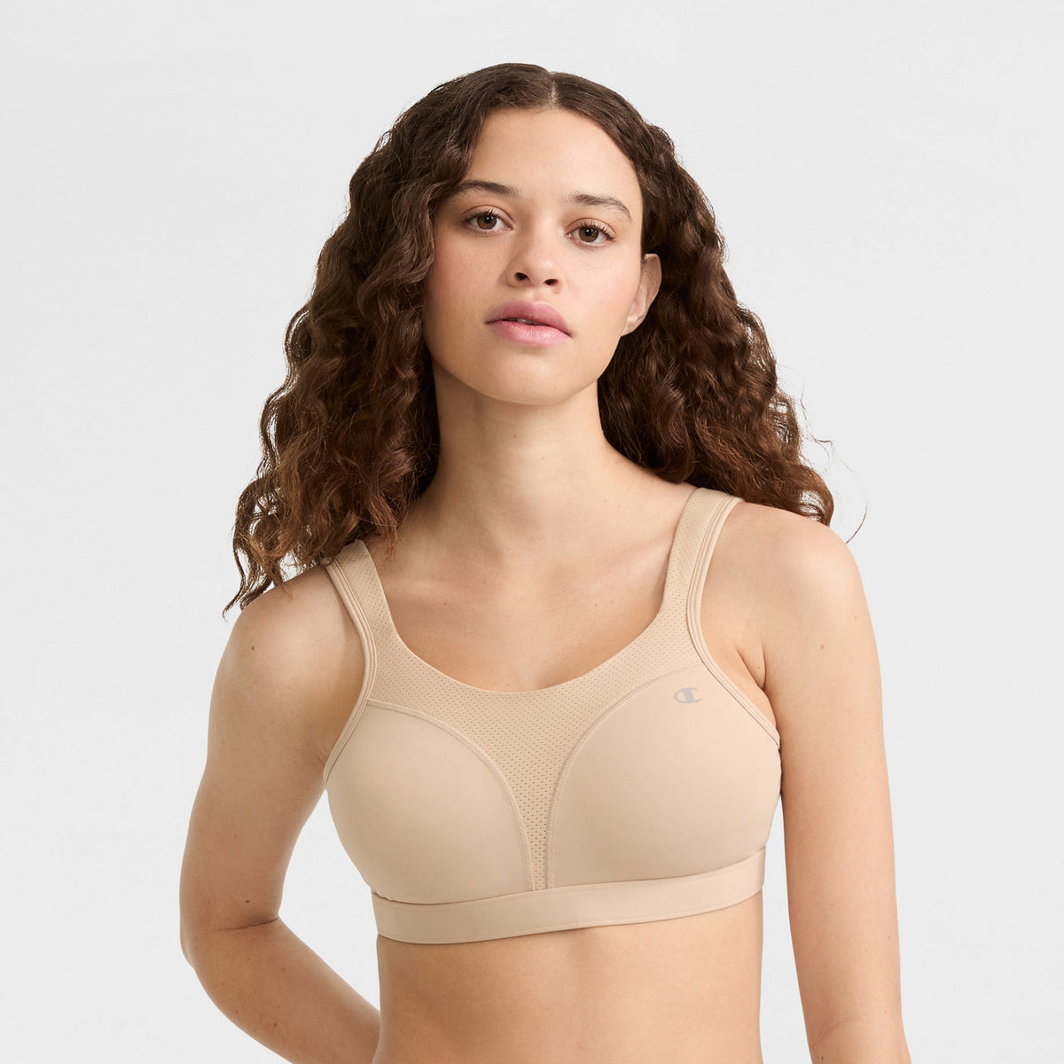 A woman with long, wavy brown hair wears the Champion Spot Comfort High-Impact Sports Bra with C Logo in beige, looking at the camera against a plain light background.
