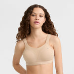 A woman with long, wavy brown hair wears the Champion Spot Comfort High-Impact Sports Bra with C Logo in beige, looking at the camera against a plain light background.