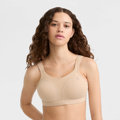 A woman with long, wavy brown hair wears the Champion Spot Comfort High-Impact Sports Bra with C Logo in beige, looking at the camera against a plain light background.